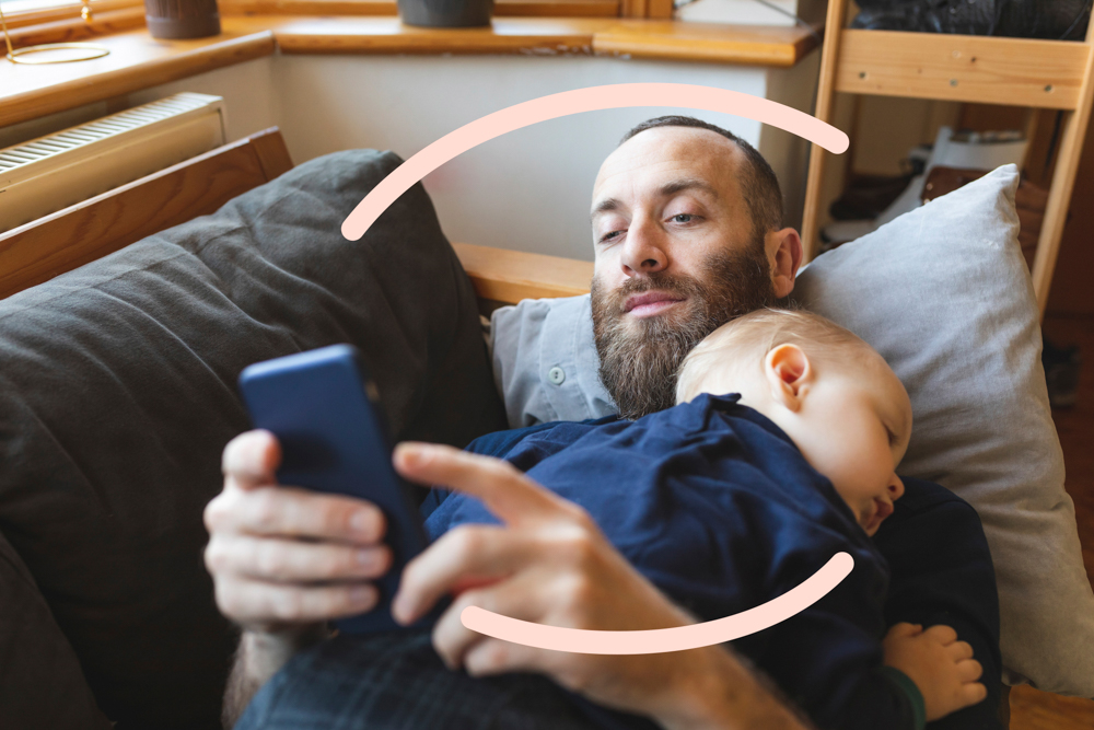 Man looking a phone whilst baby sleeps on him